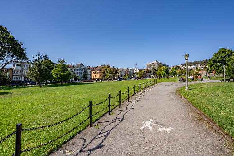 Paved walking path through Duboce Park in San Francisco’s Lower Haight, with open green lawns, surrounding Victorian buildings, and clear blue sky.