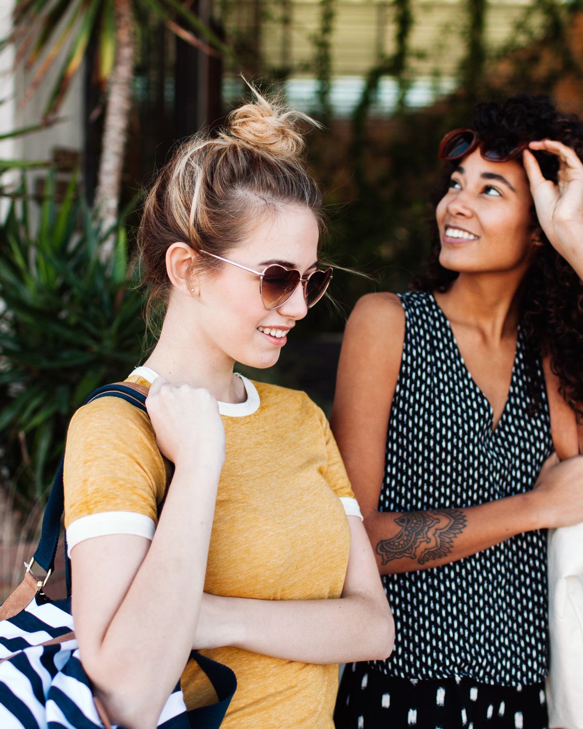 Two stylish young women enjoying a sunny day out shopping, wearing sunglasses and carrying bags, with greenery in the background.