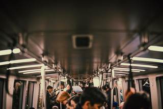 Crowded BART train interior with passengers standing and holding overhead straps, illuminated by linear ceiling lights as the train moves through the Bay Area.