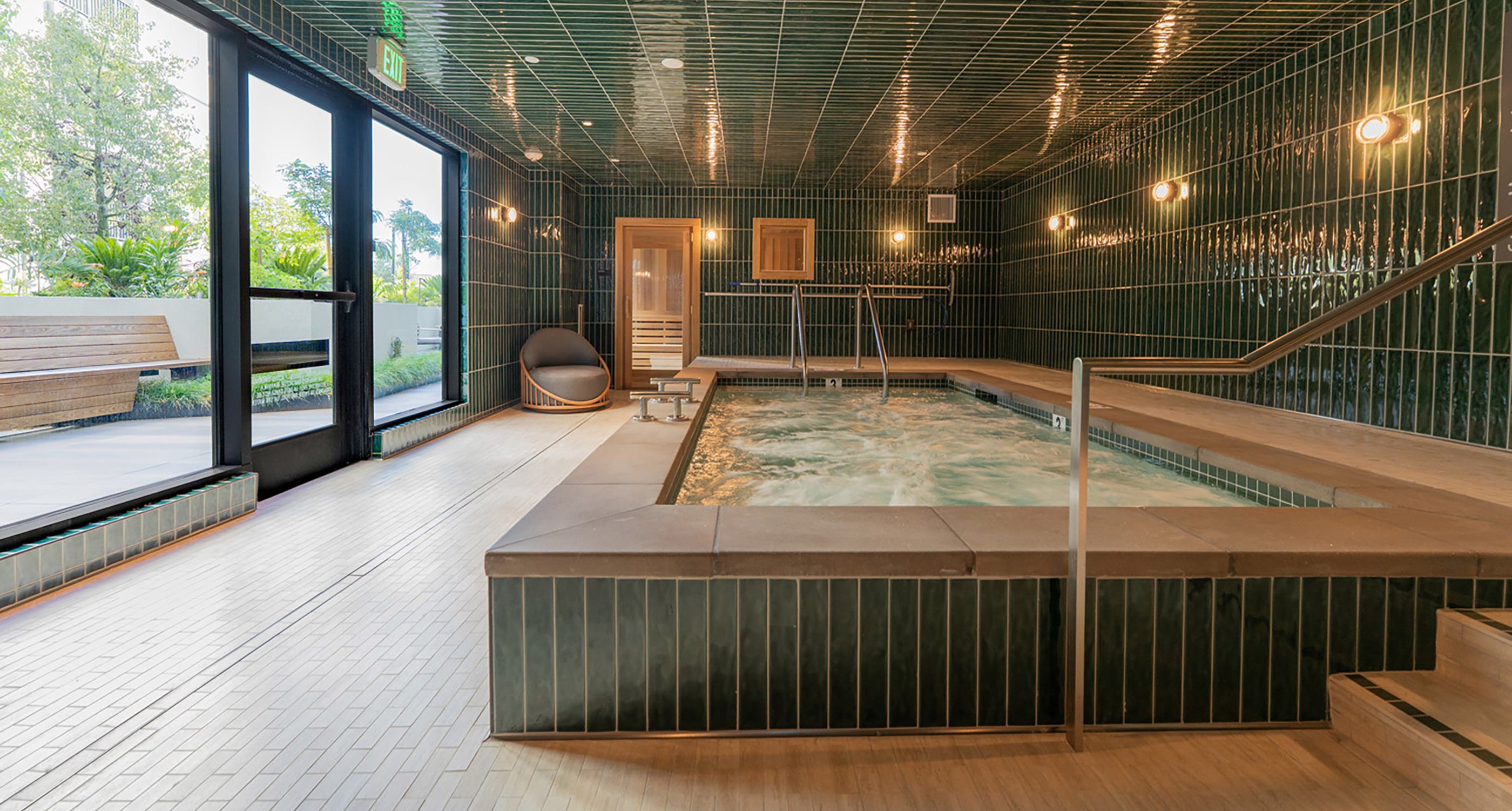 A serene indoor spa features a rectangular soaking pool framed by rich green tile walls and wood-accented sauna rooms. Floor-to-ceiling glass doors open to a tranquil outdoor lounge with blooming trees, cushioned benches, and lush landscaping.