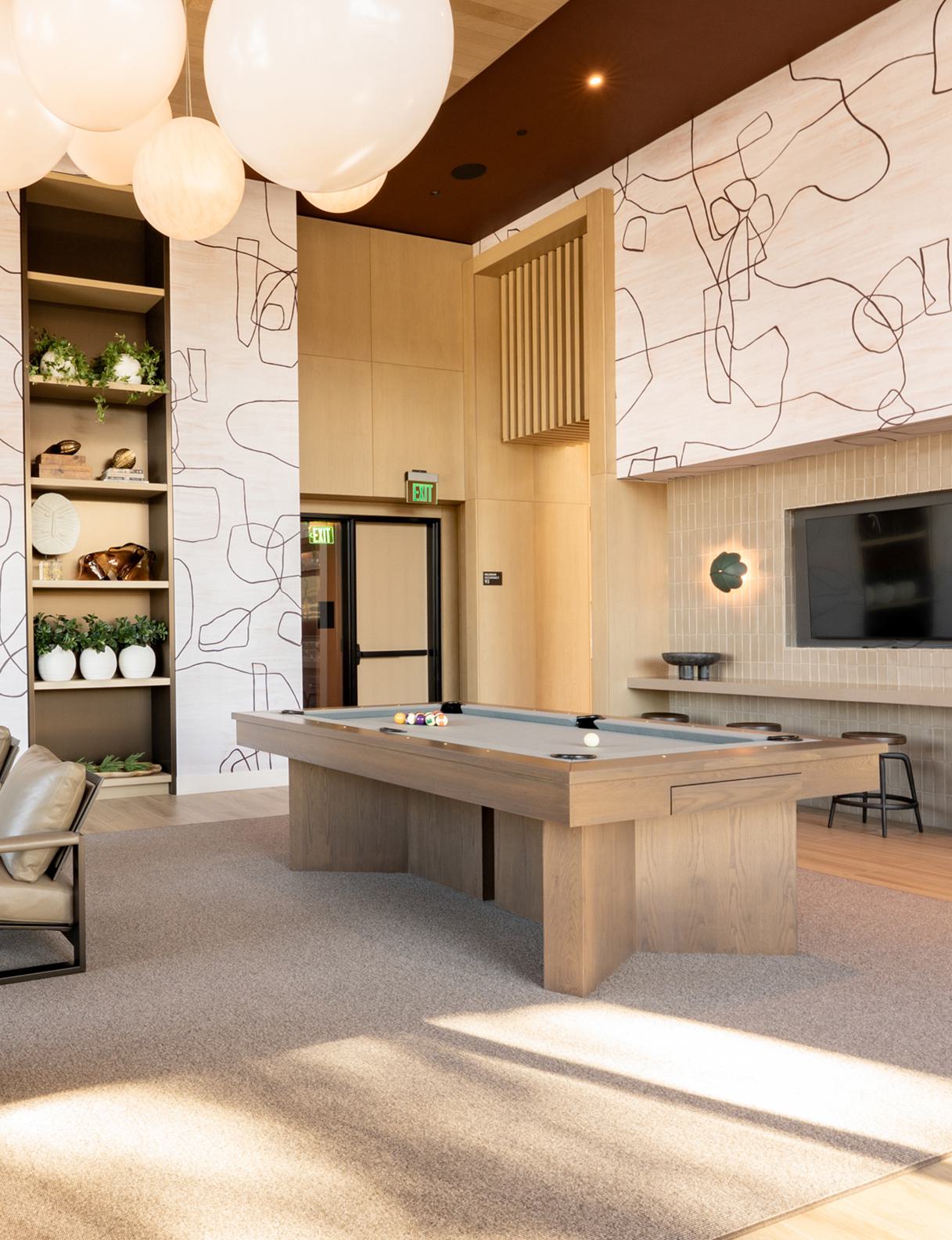 A stylish club room with sculptural globe pendant lights, textured walls, and a mix of plush seating. Floor-to-ceiling windows reveal landscaped greenery and glimpses of modern nearby buildings, creating a cozy yet connected atmosphere.