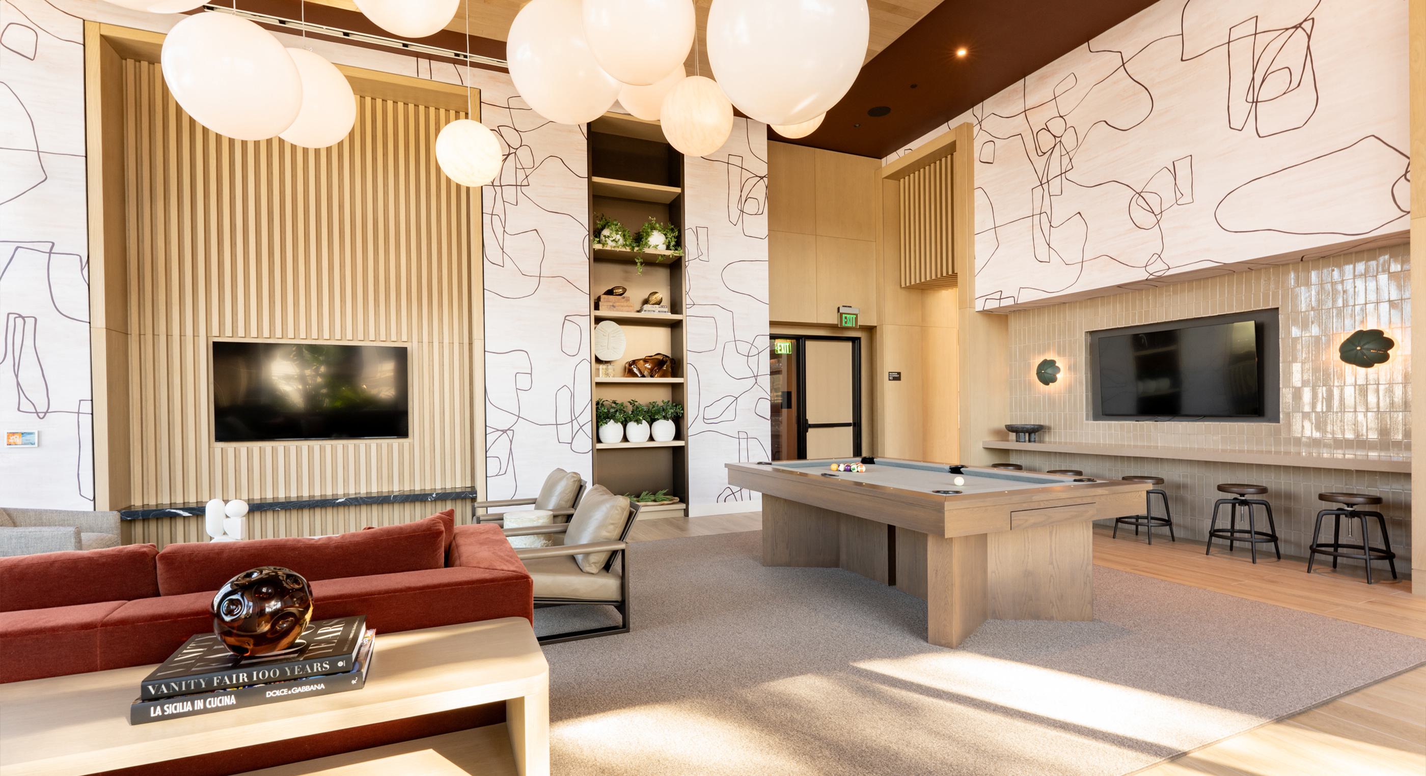 A stylish club room with sculptural globe pendant lights, textured walls, and a mix of plush seating. Floor-to-ceiling windows reveal landscaped greenery and glimpses of modern nearby buildings, creating a cozy yet connected atmosphere.