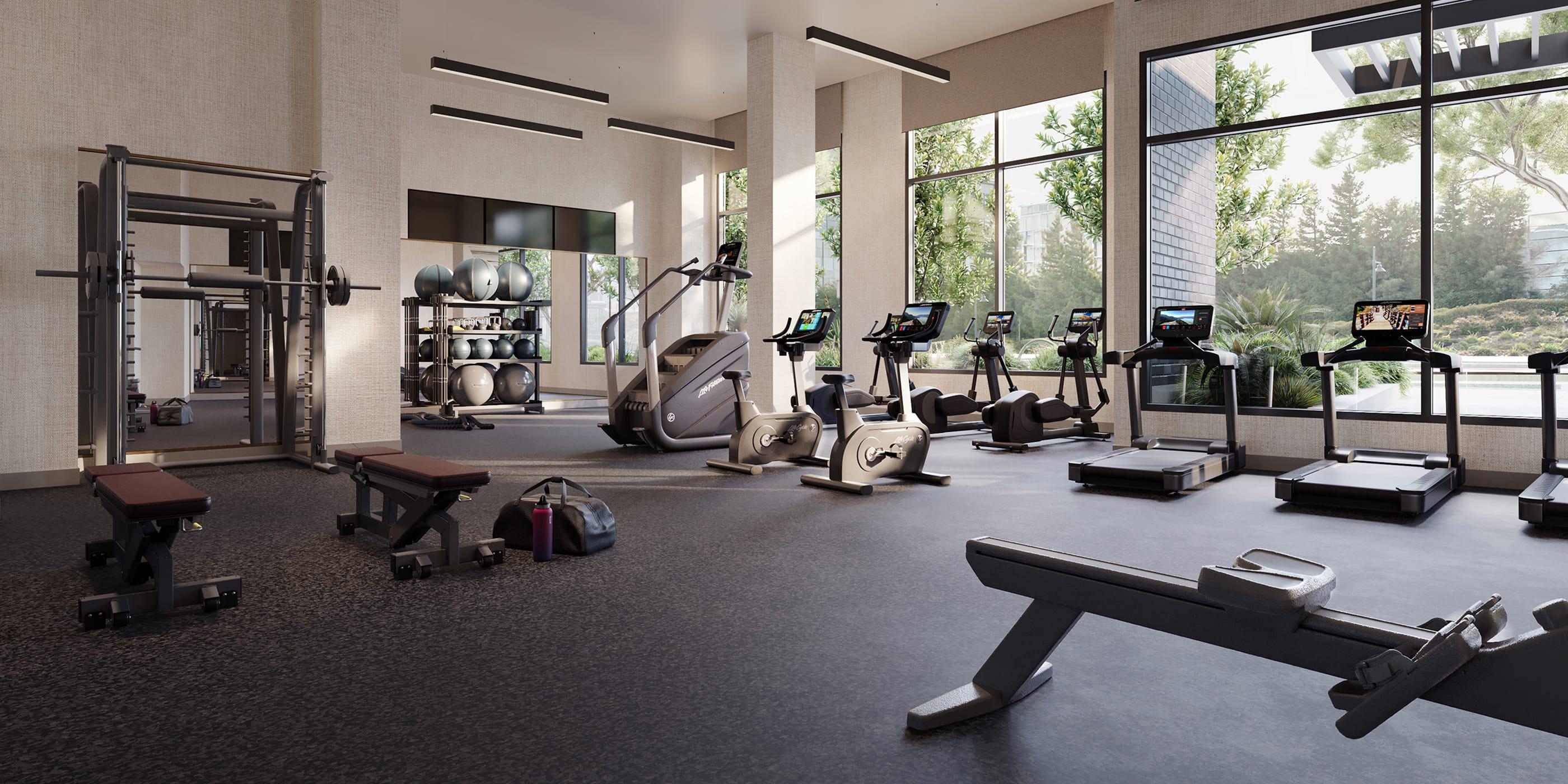 Modern fitness center with large windows overlooking greenery, equipped with treadmills, stationary bikes, weight benches, resistance machines, and racks of exercise balls.