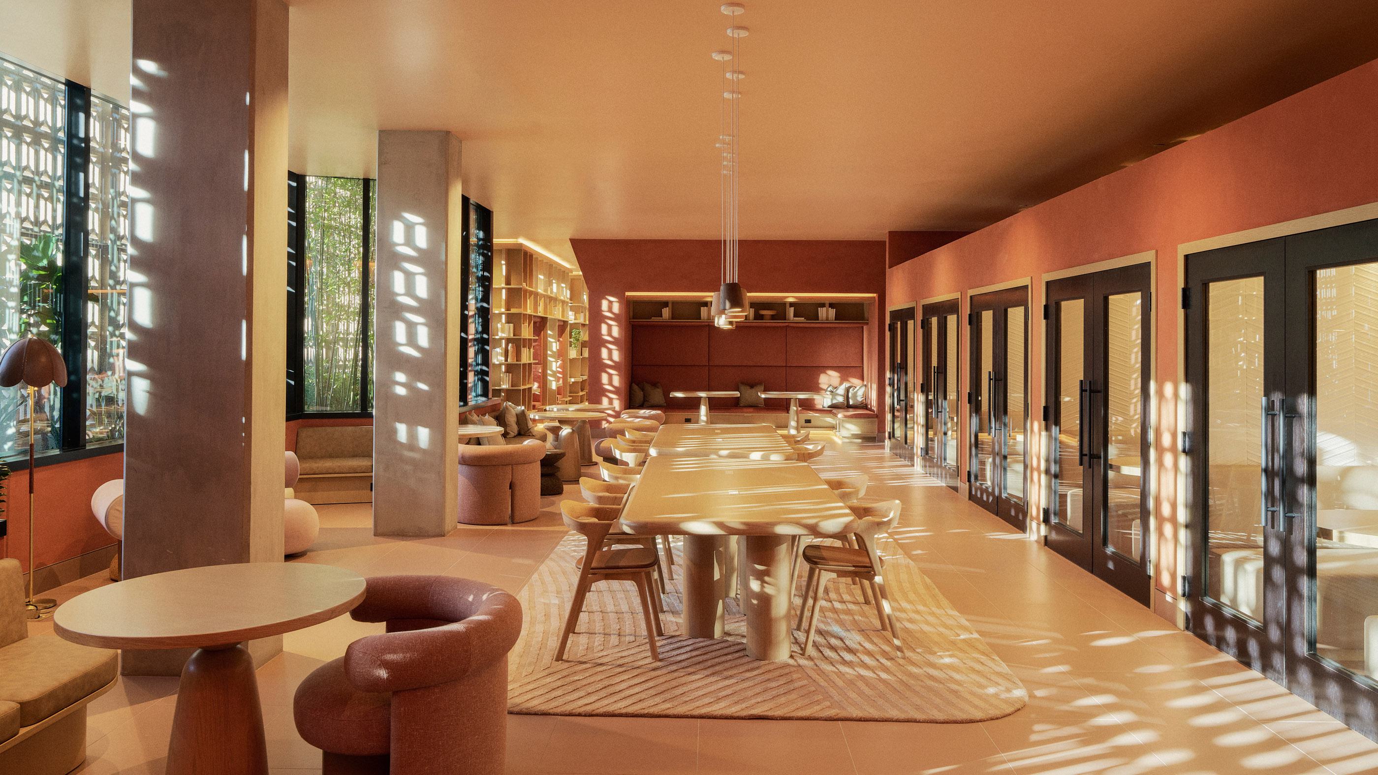 Co-working lounge with earthy tones, cozy seating, expansive bookshelves, large shared tables, and lush greenery visible through oversized windows.