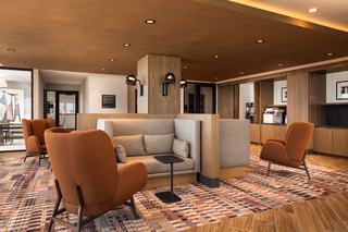 Modern coworking lounge featuring a mix of semi-private seating nooks and mid-century-inspired armchairs in warm terracotta upholstery. The space includes a patterned area rug, wood-paneled ceiling, and built-in cabinetry with office equipment, creating a refined, functional environment for work or casual meetings.