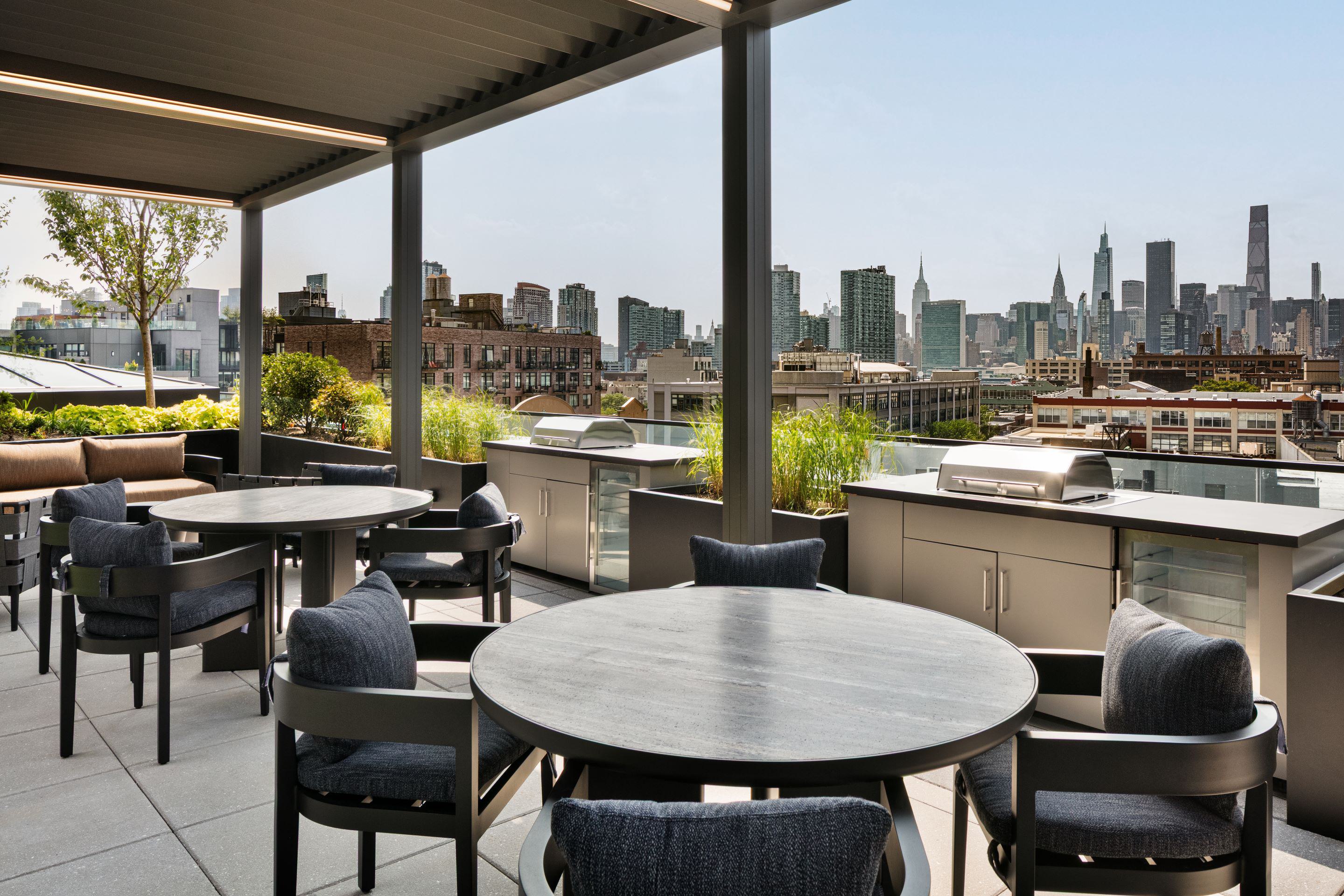 Outdoor terrace dining area at Lumen in Long Island City, with modern pergolas, communal tables, and lounge seating arranged among greenery, offering views of the Manhattan skyline in the background.