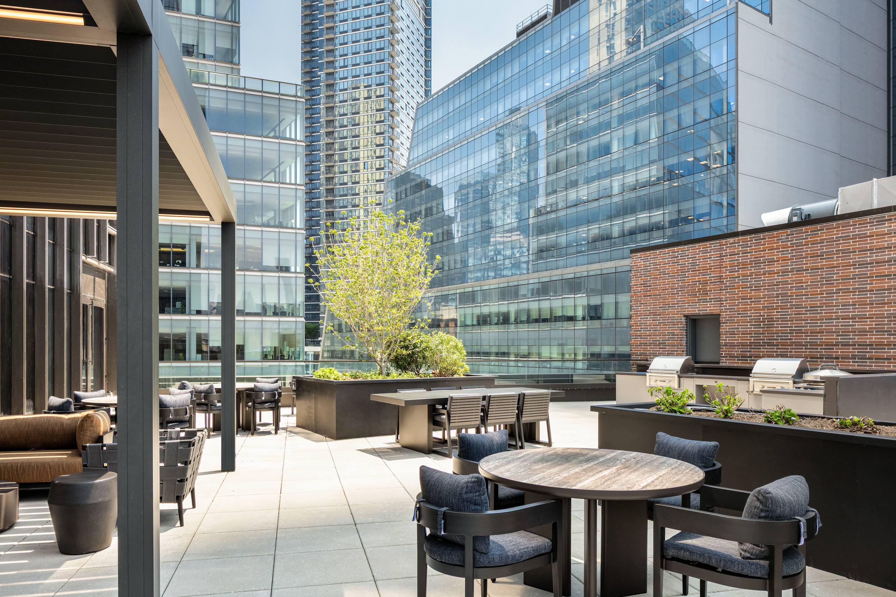 Outdoor grilling station on the terrace at Lumen in Long Island City, featuring built-in stainless steel grills, prep counters, shaded dining tables, and surrounding planters, with high-rise buildings enclosing the courtyard-style space.