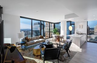  A modern open-concept living room and kitchen in a high-rise apartment at Atlas in downtown Oakland. The space features floor-to-ceiling corner windows that wrap around the dining and living areas, offering expansive views of the Tribune Tower’s iconic signage, nearby office buildings, and the distant San Francisco Bay. The interior includes light wood flooring, a white quartz waterfall kitchen island, mid-century inspired furniture, and abundant natural light flooding the space. A round dining table with curved chairs and large indoor plants add warmth to the contemporary setting.