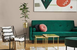 Modern living room with a green velvet sofa, round pink and navy pillows, gold nesting tables, a potted plant in a brass stand, and a black wire chair with a striped throw blanket.