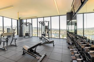 High-rise fitness center at Atlas Apartments in Oakland featuring weight machines, free weights, and floor-to-ceiling windows with panoramic views of Lake Merritt and the East Bay.