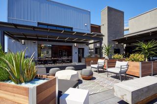Outdoor rooftop lounge at Fourth Street East featuring modern seating areas, patterned rugs, concrete and wood planter boxes with succulents, a fire pit, and covered dining space with a long communal table, set against a clear blue sky.
