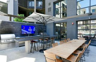 Modern outdoor grilling and dining area at Fourth Street East, featuring stainless steel gas grills, a wall-mounted TV, bar seating with umbrellas, and a long wooden dining table with chairs, set against a courtyard backdrop.
