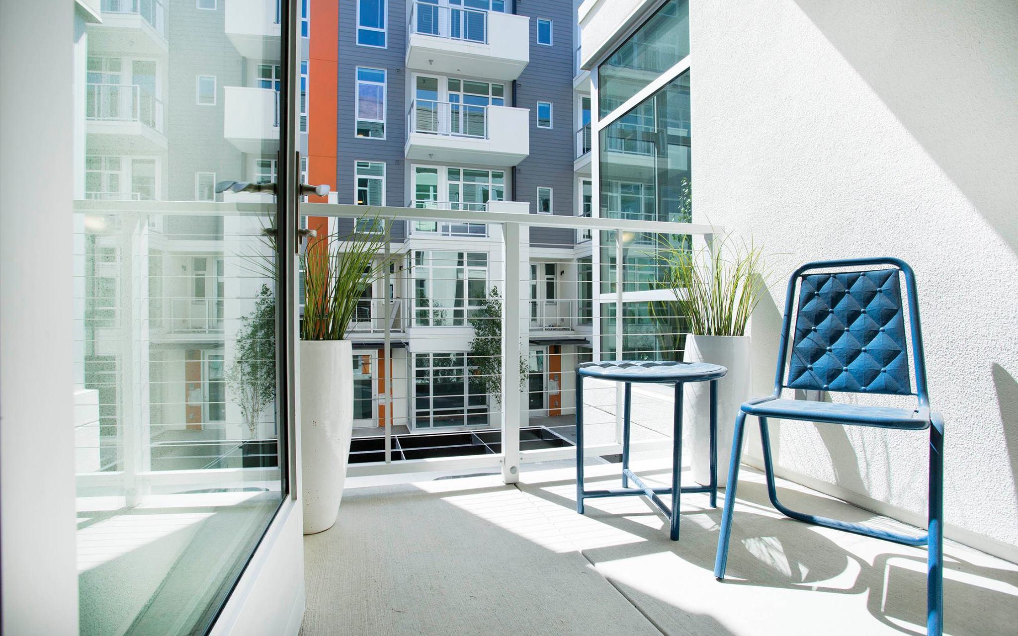 Private balcony at Alchemy with modern blue chair, small table, and tall planters, overlooking contemporary apartment buildings with glass railings.