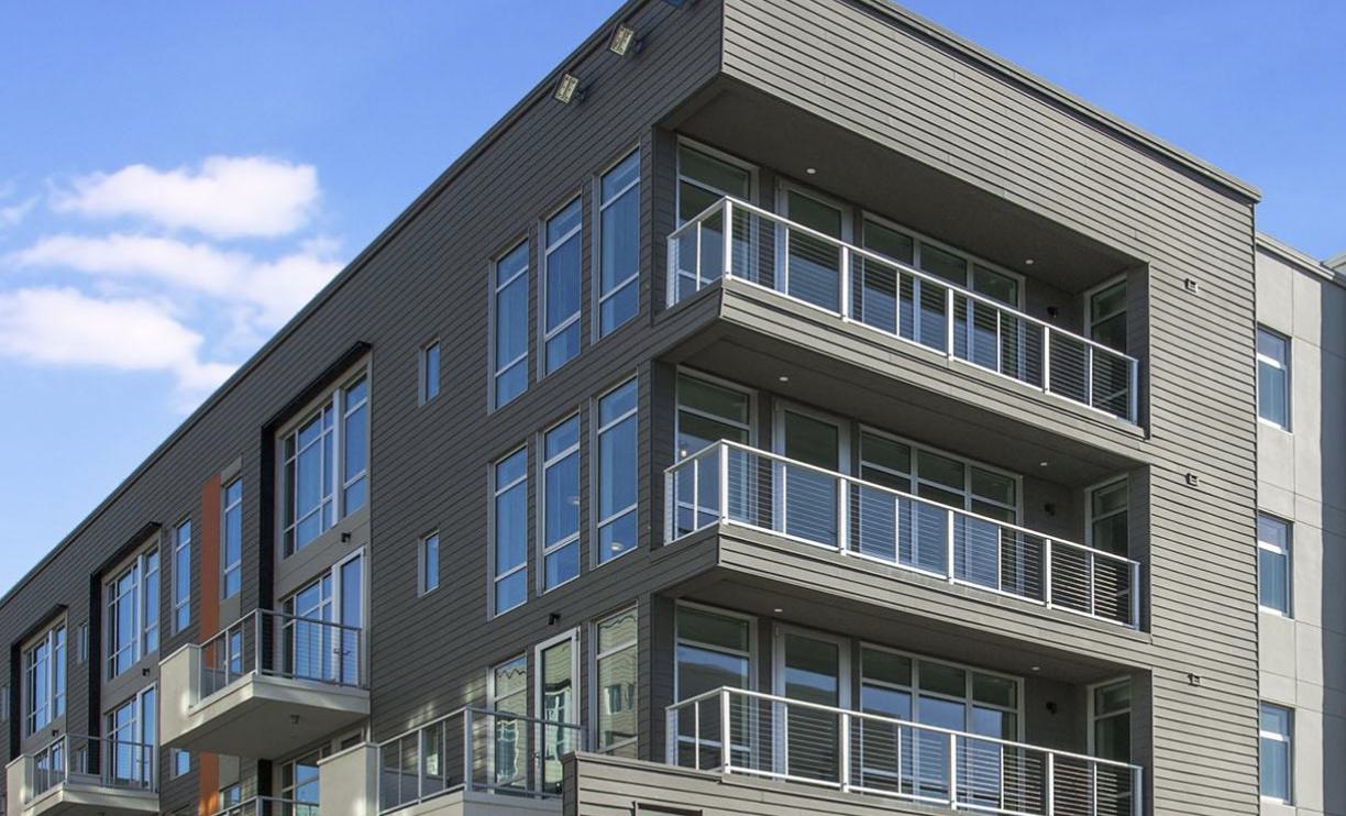 A modern apartment building features large windows and multiple private balconies with sleek metal railings. The clean lines, neutral siding, and minimalist design create a contemporary urban look under a clear blue sky.