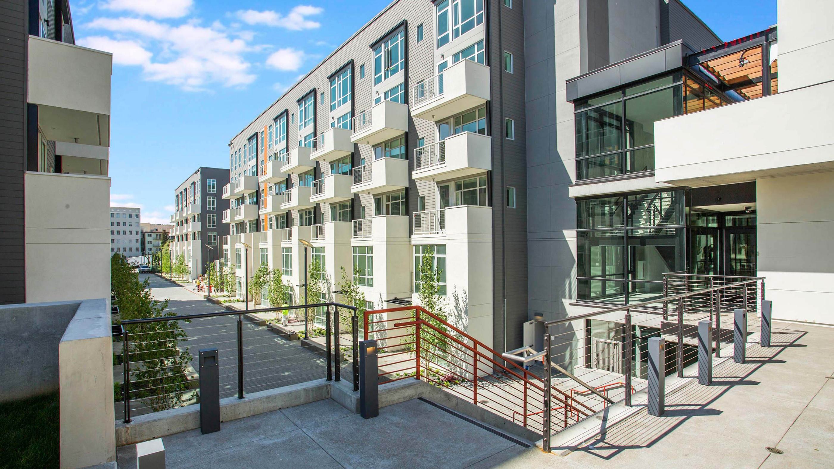 Modern apartment buildings line a landscaped breezeway with young trees and private balconies. The design features breezeway-level access to some residences, with glass stairwells, metal railings, and clean concrete walkways highlighting the contemporary urban aesthetic.