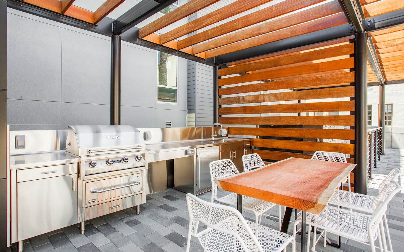 Modern outdoor BBQ area at Alchemy featuring a sleek stainless steel grill and prep station, shaded by a wood-slatted pergola with dining table and chairs.