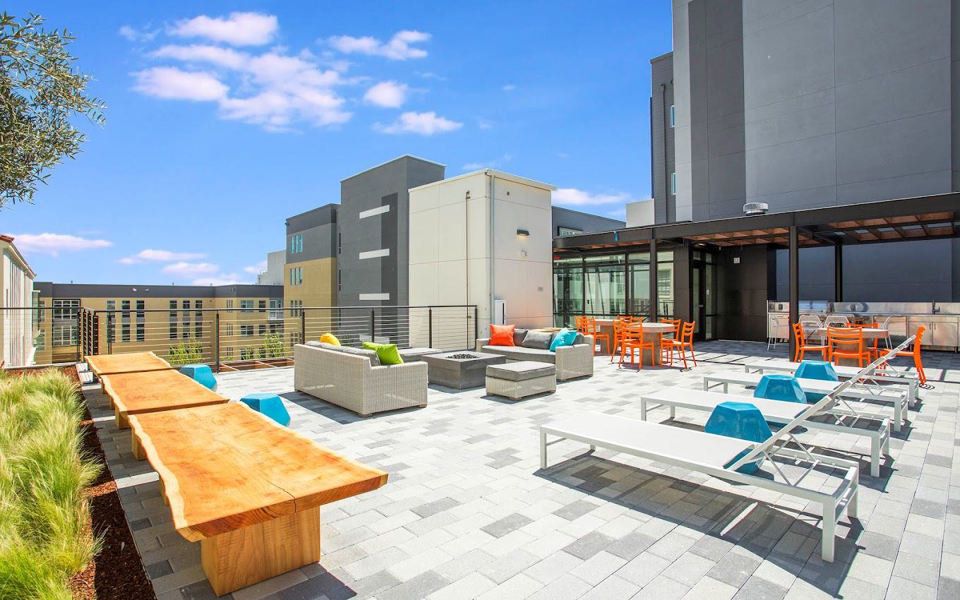 Alchemy Apartments deck in San Francisco featuring cushioned lounge seating, wooden benches, sun loungers, bright orange dining chairs, and a built-in grilling station under a modern pergola.