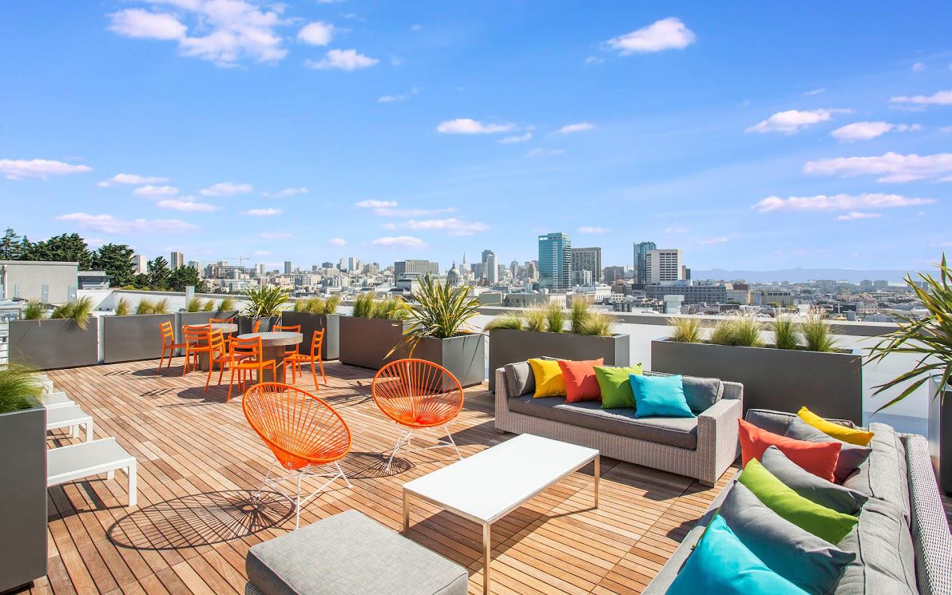 Colorful rooftop lounge at Alchemy Apartments in Hayes Valley, with wooden decking, vibrant orange chairs, and cushioned seating overlooking the San Francisco skyline under a bright blue sky