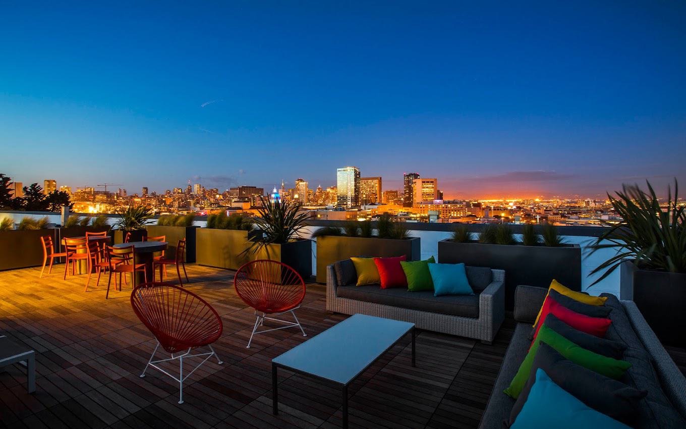 Evening view of a rooftop terrace featuring Acapulco-style chairs, colorful cushioned sofas, and dining tables overlooking the illuminated San Francisco skyline.