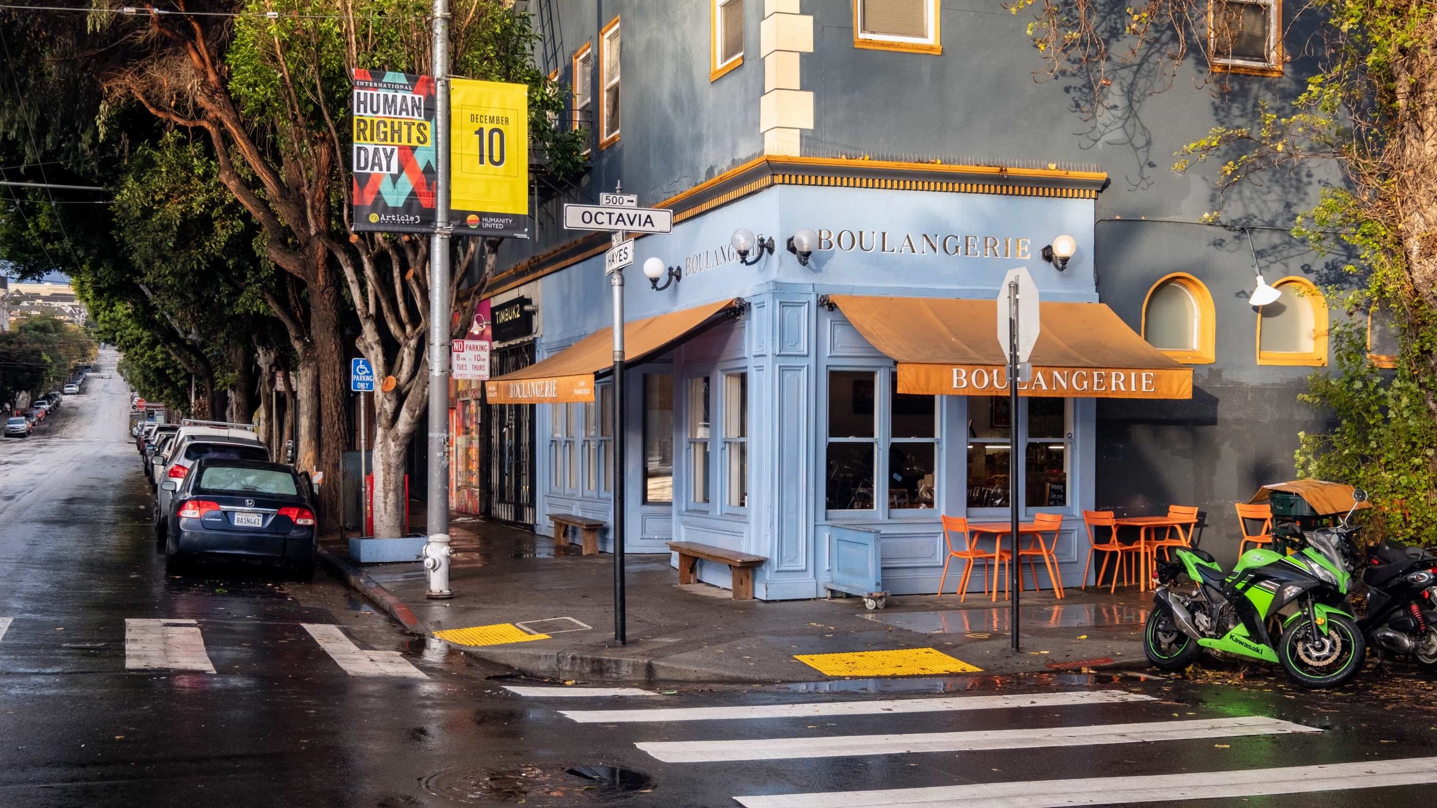 Corner view of La Boulangerie on Hayes and Octavia in San Francisco, with a blue facade, orange awnings and chairs, and tree-lined streets after rain.