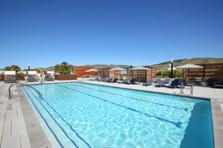 Rooftop lap pool at The Asher with three swim lanes, surrounded by loungers, shaded cabanas, and views of the rolling East Bay hills under a clear blue sky.