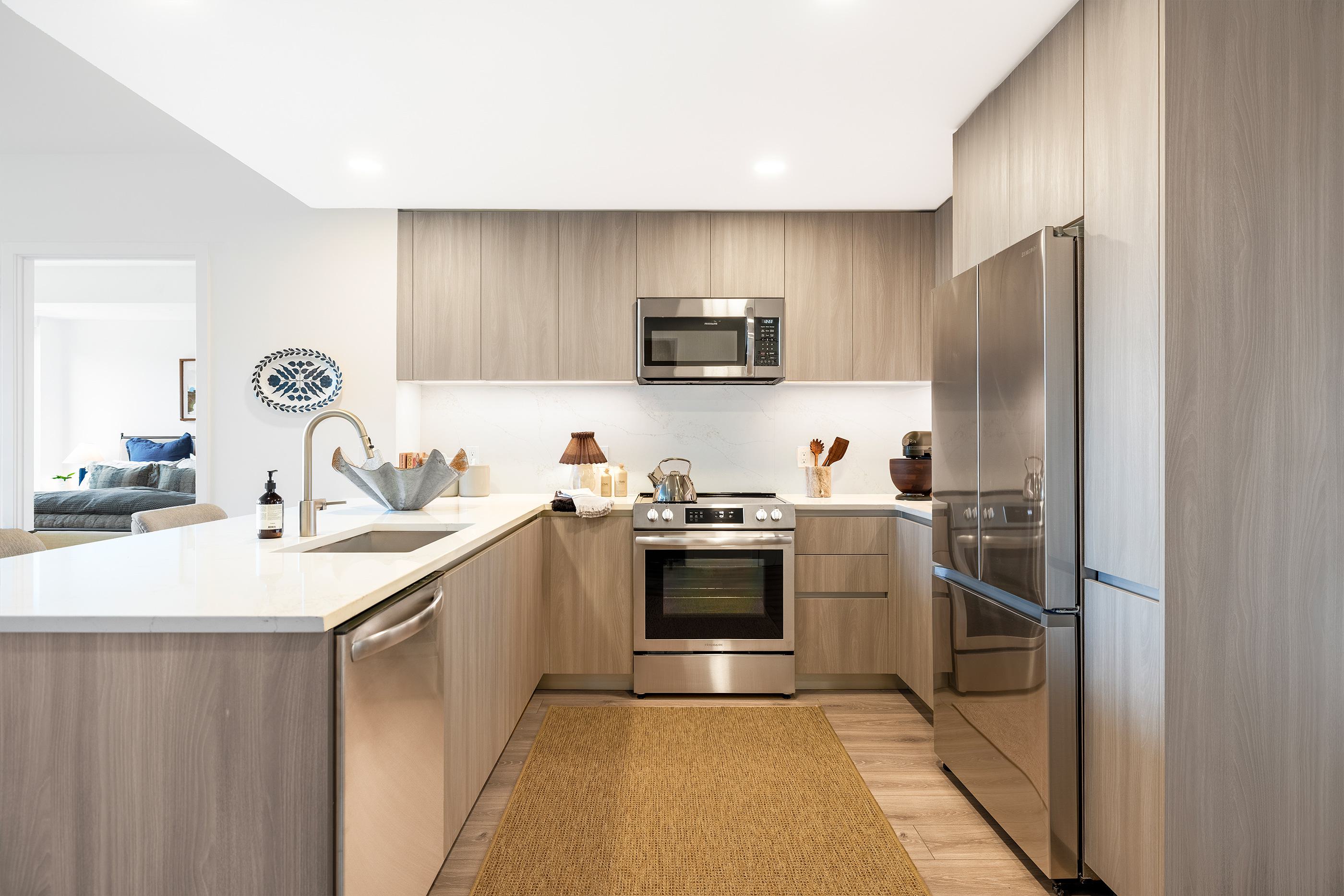 Modern kitchen at Zephyr in Woodley Park, DC, featuring warm wood cabinetry, sleek Bosch stainless steel appliances, quartz countertops, a marble-look backsplash, and a peninsula with upholstered bar seating.