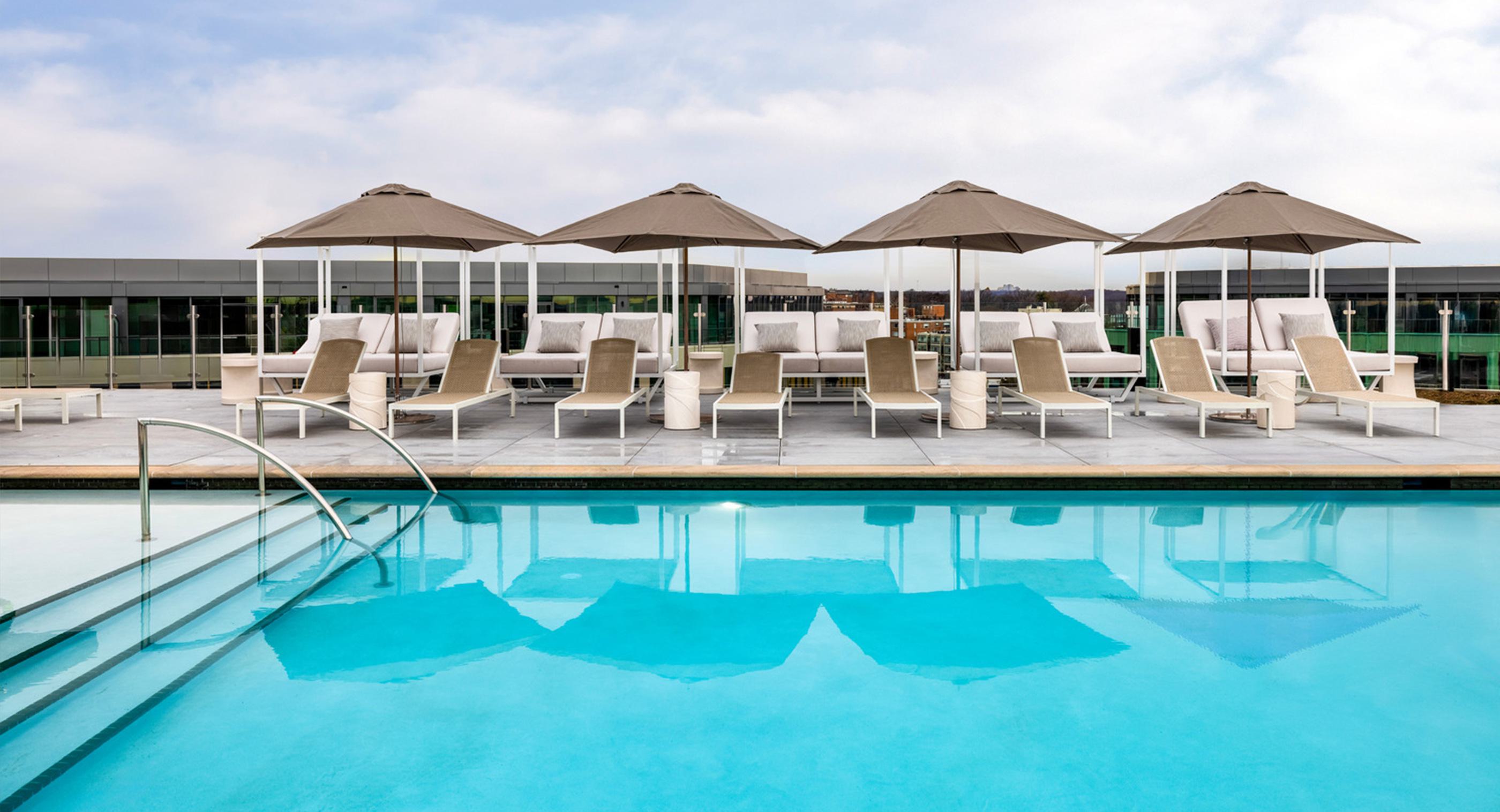 Rooftop pool at Zephyr in Woodley Park, DC, with in-water loungers, shaded cabanas, and panoramic city views, including the Washington Monument in the distance at sunset.