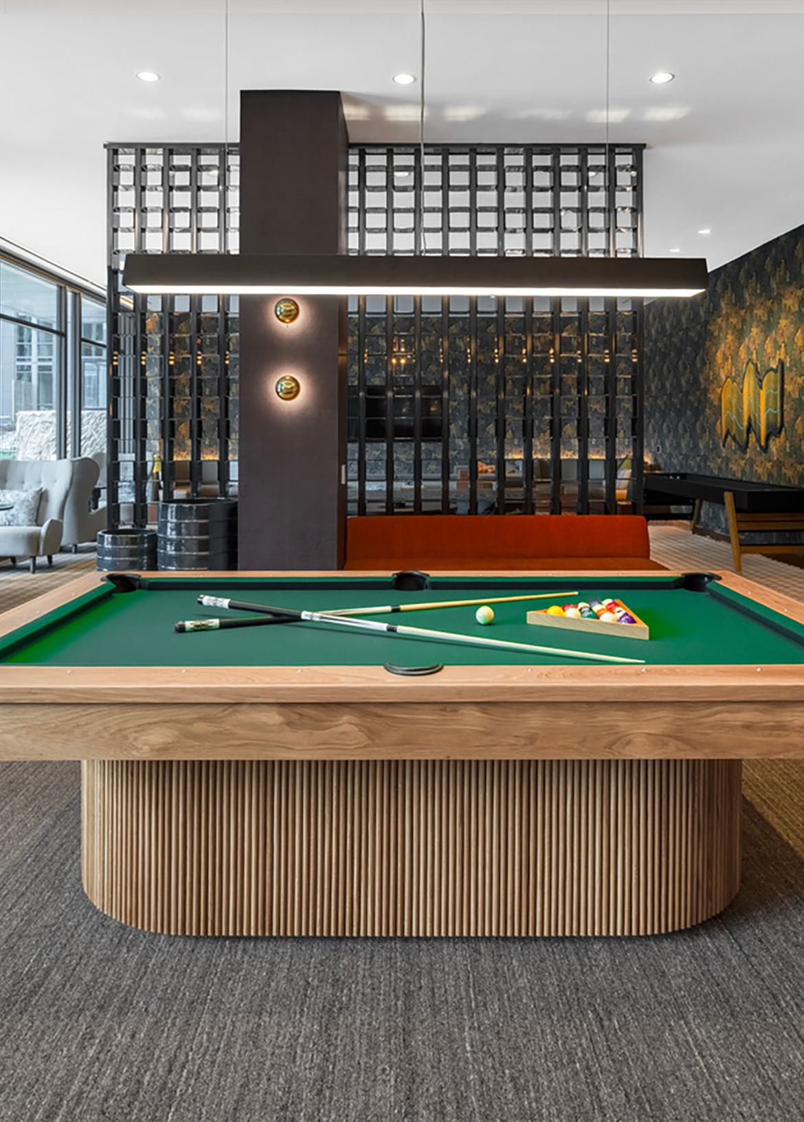 Game lounge at Zephyr in Woodley Park, DC, featuring a pool table, foosball table, bar-style seating, and cozy booths, with warm lighting and floor-to-ceiling windows overlooking landscaped outdoor areas.