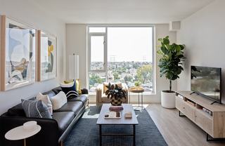 Living room with floor-to-ceiling window overlooking a low-rise residential neighborhood in Los Angeles’ West Adams area, featuring tree-lined streets, rooftops, and utility towers in the distance under a bright, hazy sky.