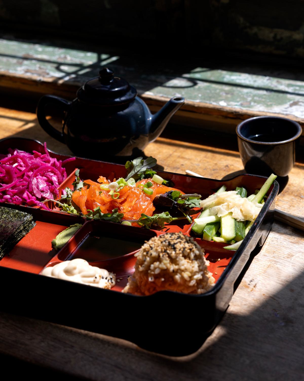Close-up of a vibrant bento box with fresh salmon, pickled vegetables, cucumber, and a sesame rice ball, paired with a teapot and cup, representing the diverse dining options in Woodley Park, DC.
