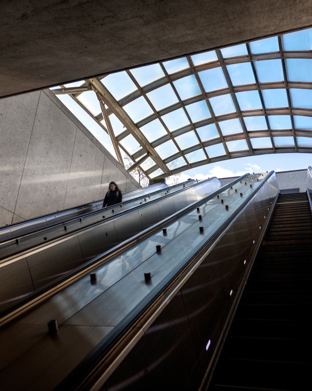 Escalator ascending to the street level at the Woodley Park Metro station in Washington, DC, framed by the station's glass canopy roof with natural light streaming in.
