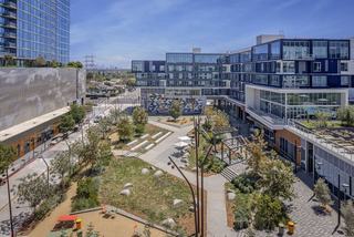 Aerial view of the Zócalo at VOX in West Adams, Los Angeles — a one-acre urban park with walking paths, landscaped lawns, seating areas, and shaded structures, surrounded by modern mid-rise apartment buildings and retail spaces.
