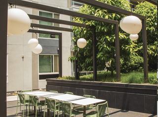 Outdoor dining space at VOX with a long white table and green woven chairs under a modern pergola adorned with spherical pendant lights, surrounded by lush greenery.