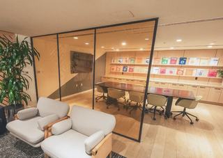 Glass-walled conference room with a long black meeting table, green swivel chairs, and a wall of colorful travel-themed books above light wood cabinetry, viewed from an adjacent lounge area with two cushioned armchairs and a potted plant.