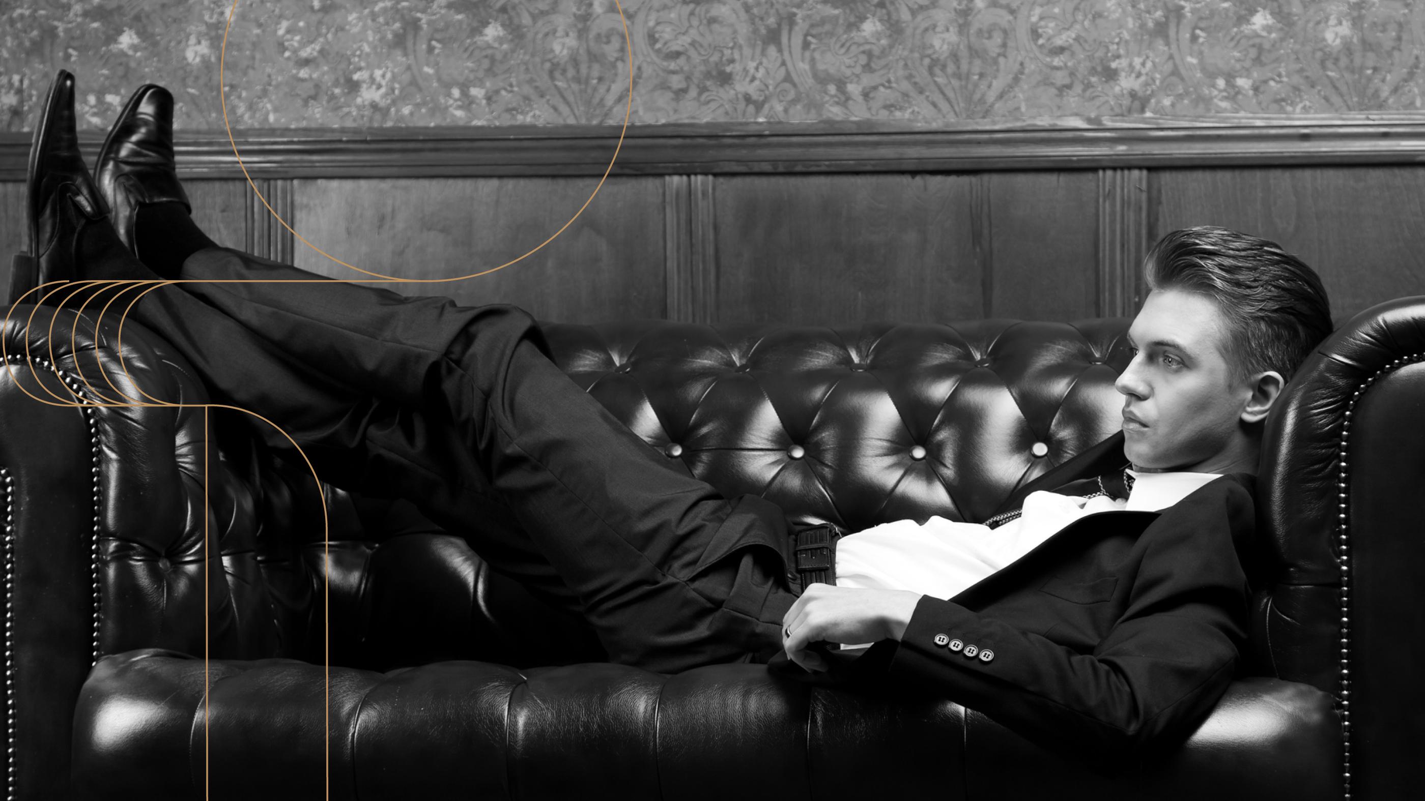 Black-and-white photo of a man in a suit lounging on a tufted leather sofa in a refined, vintage-style setting.