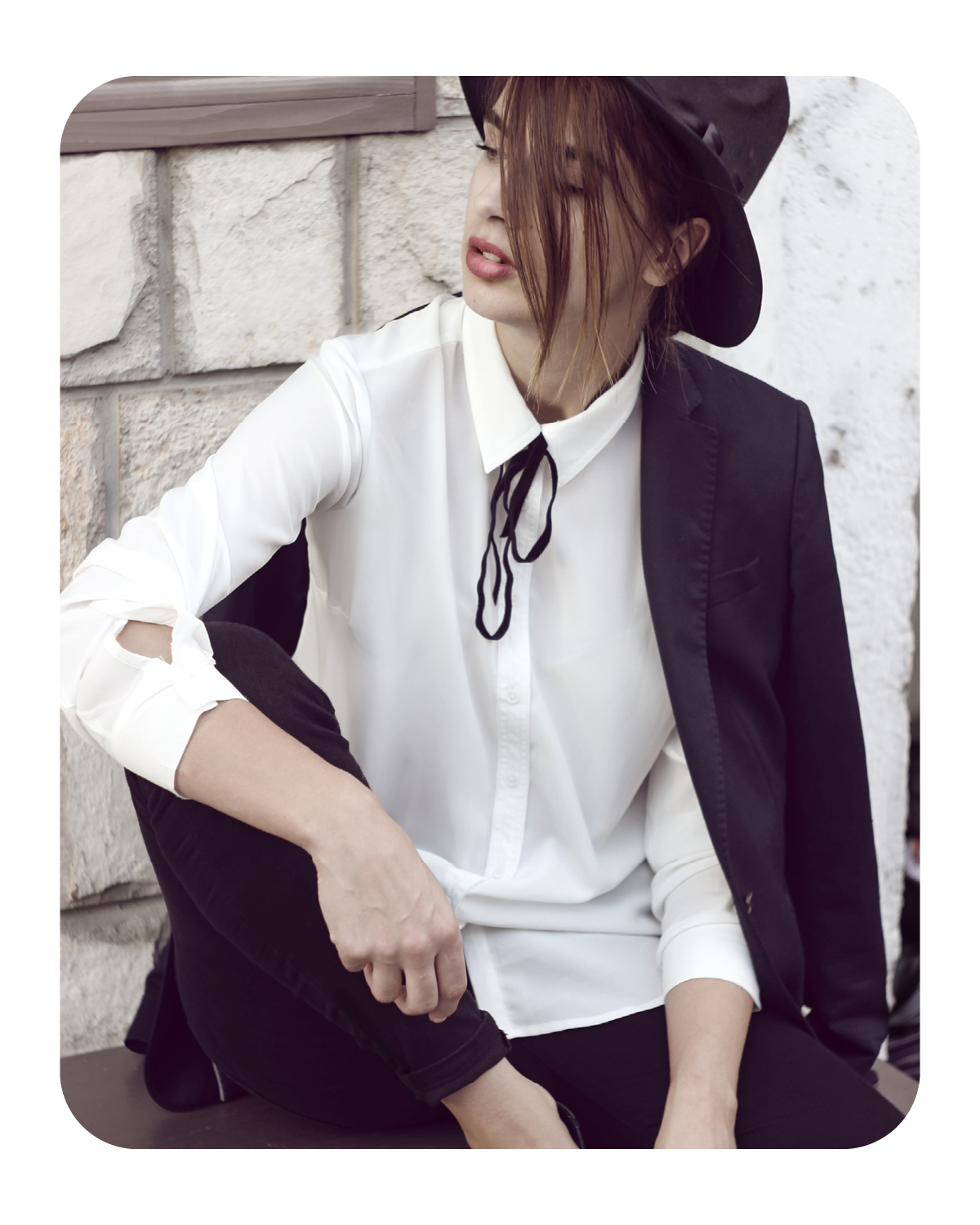 Stylish woman in a black hat and blazer sitting casually against a stone wall.