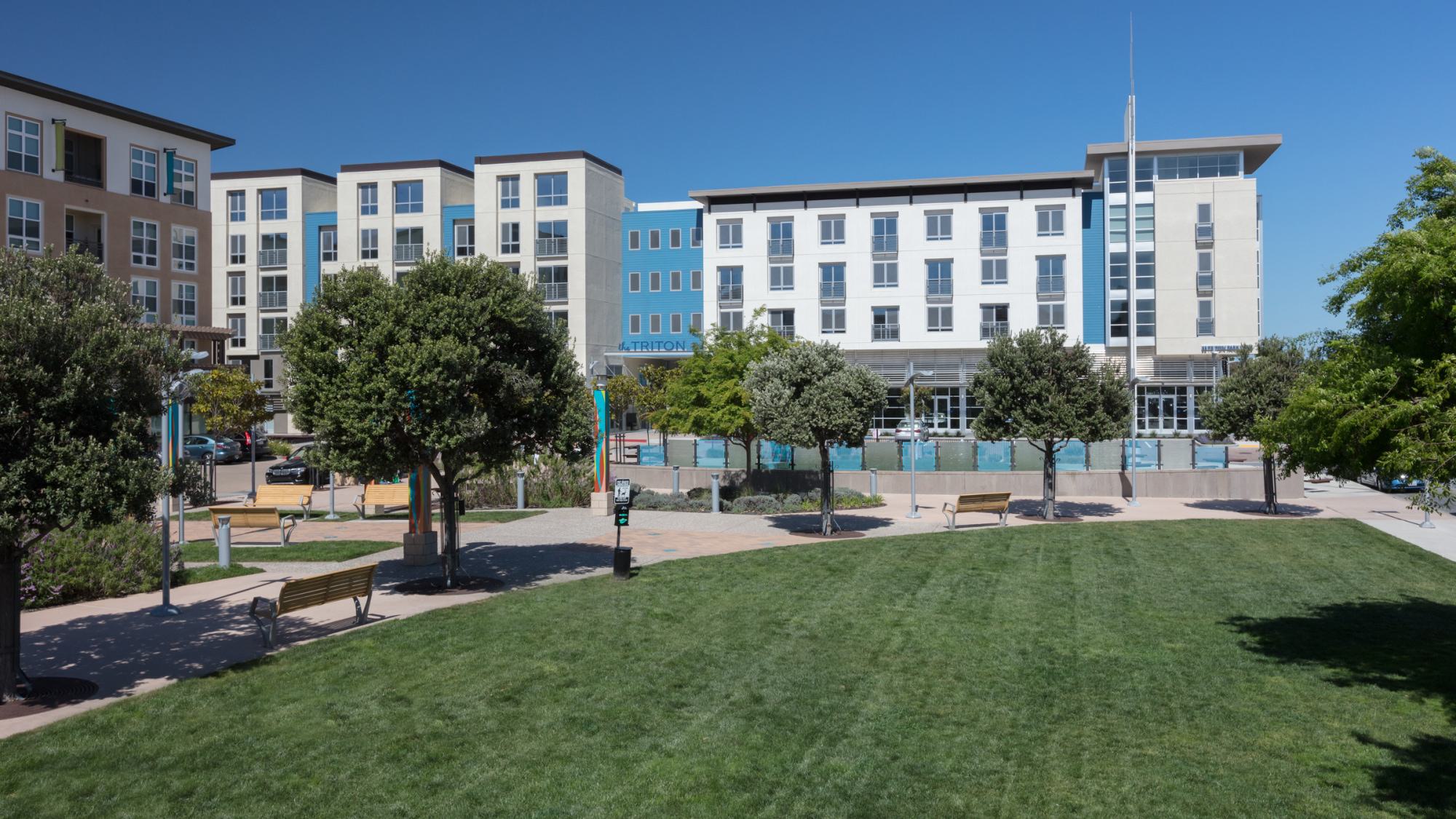 View of The Triton apartments in Foster City with landscaped green space, walking paths, benches, and shaded trees in the foreground.