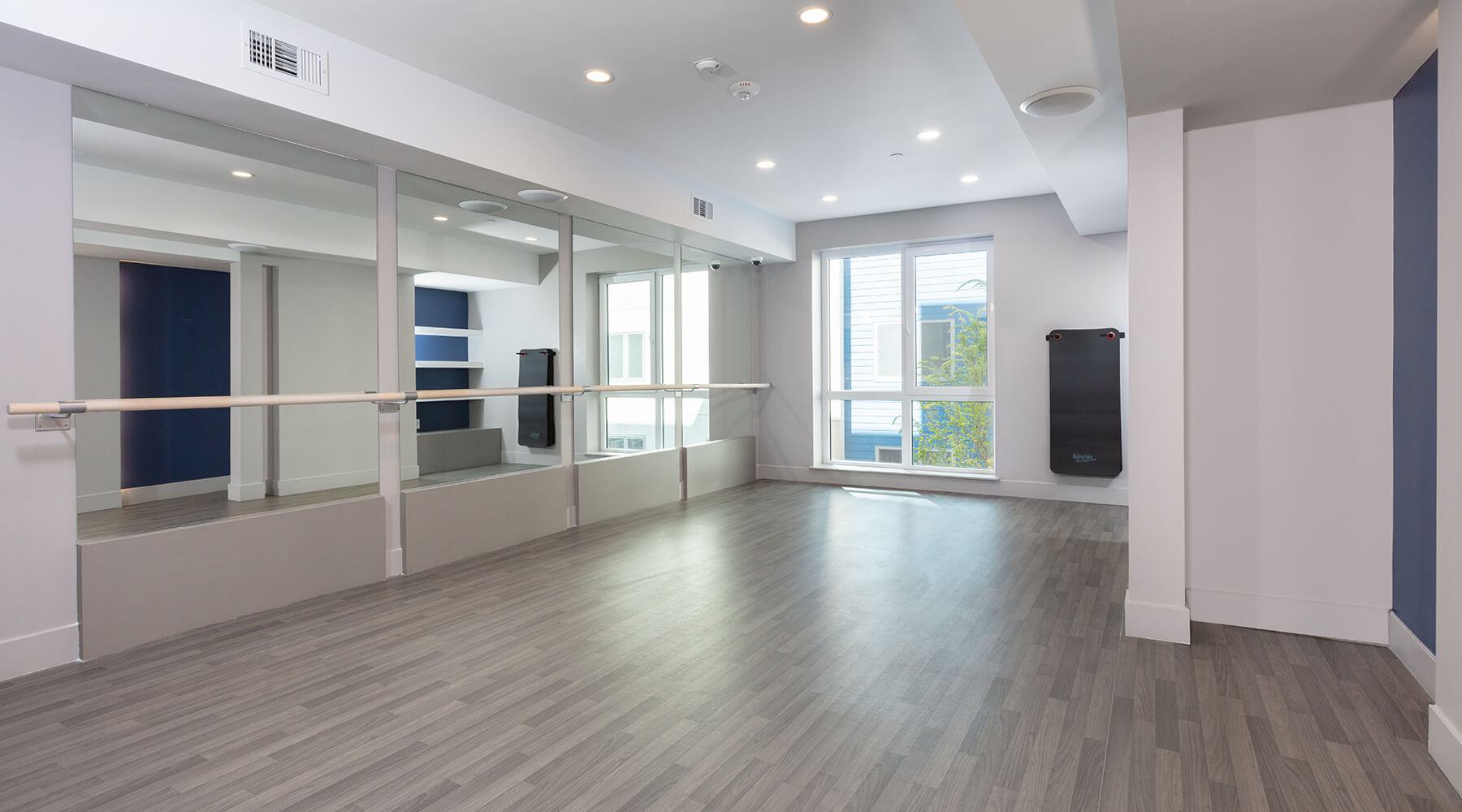 yoga and barre studio at The Triton with mirrored walls, ballet barre, wood-style flooring, and large window bringing in natural light.