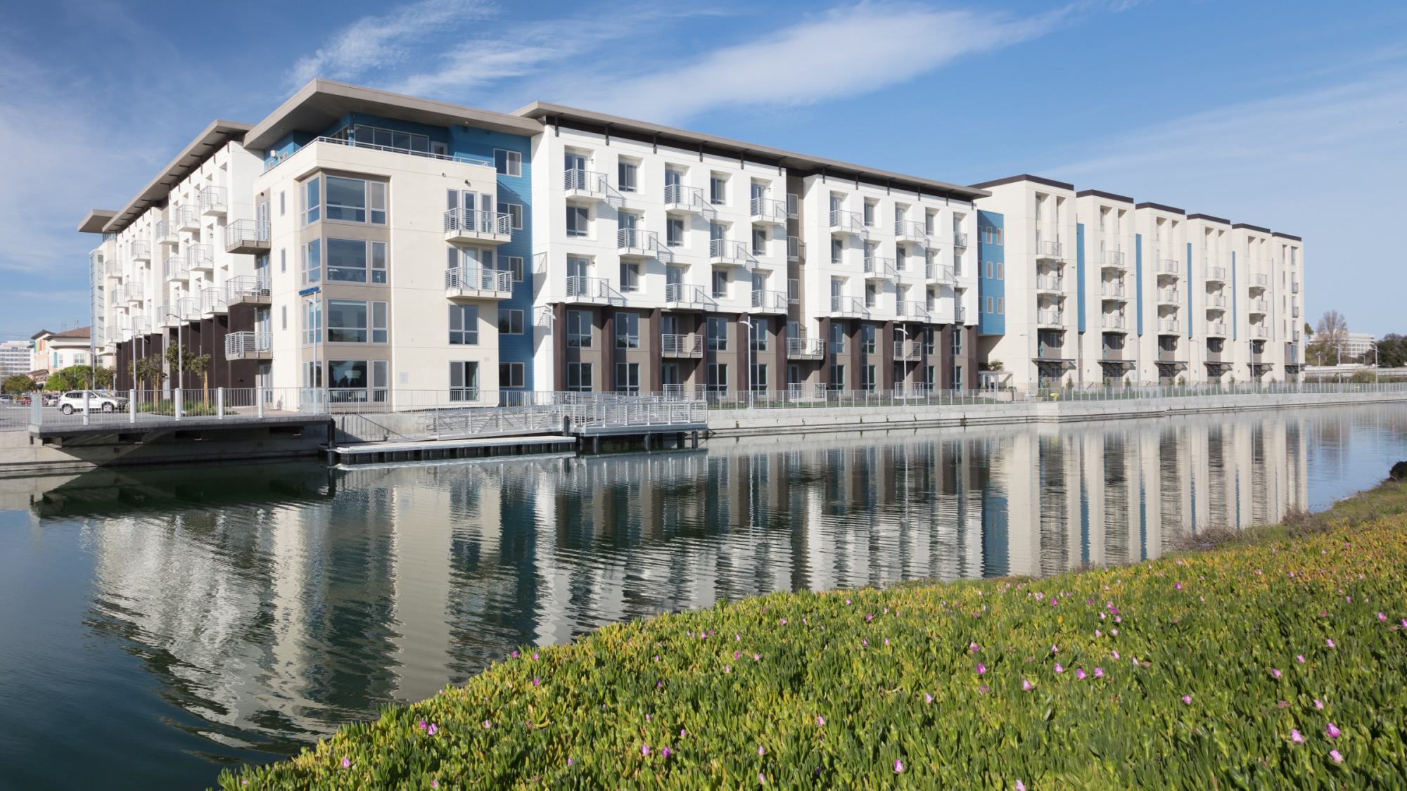 The Triton apartments in Foster City with balconies overlooking the lagoon, reflected in the calm water with greenery in the foreground.