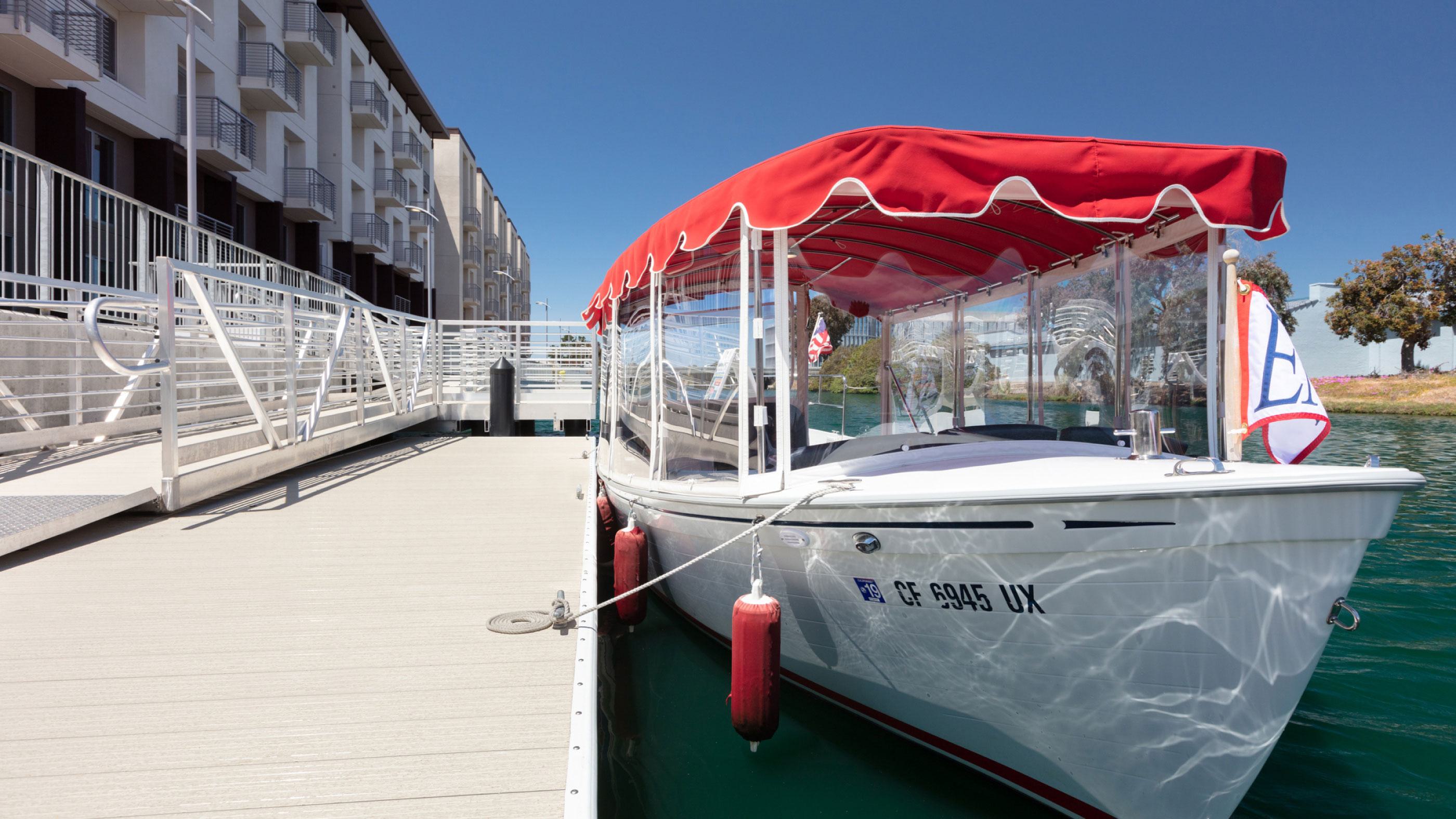 Boat docked at The Triton’s private waterfront launch in Foster City, with red canopy and apartment building visible along the lagoon.