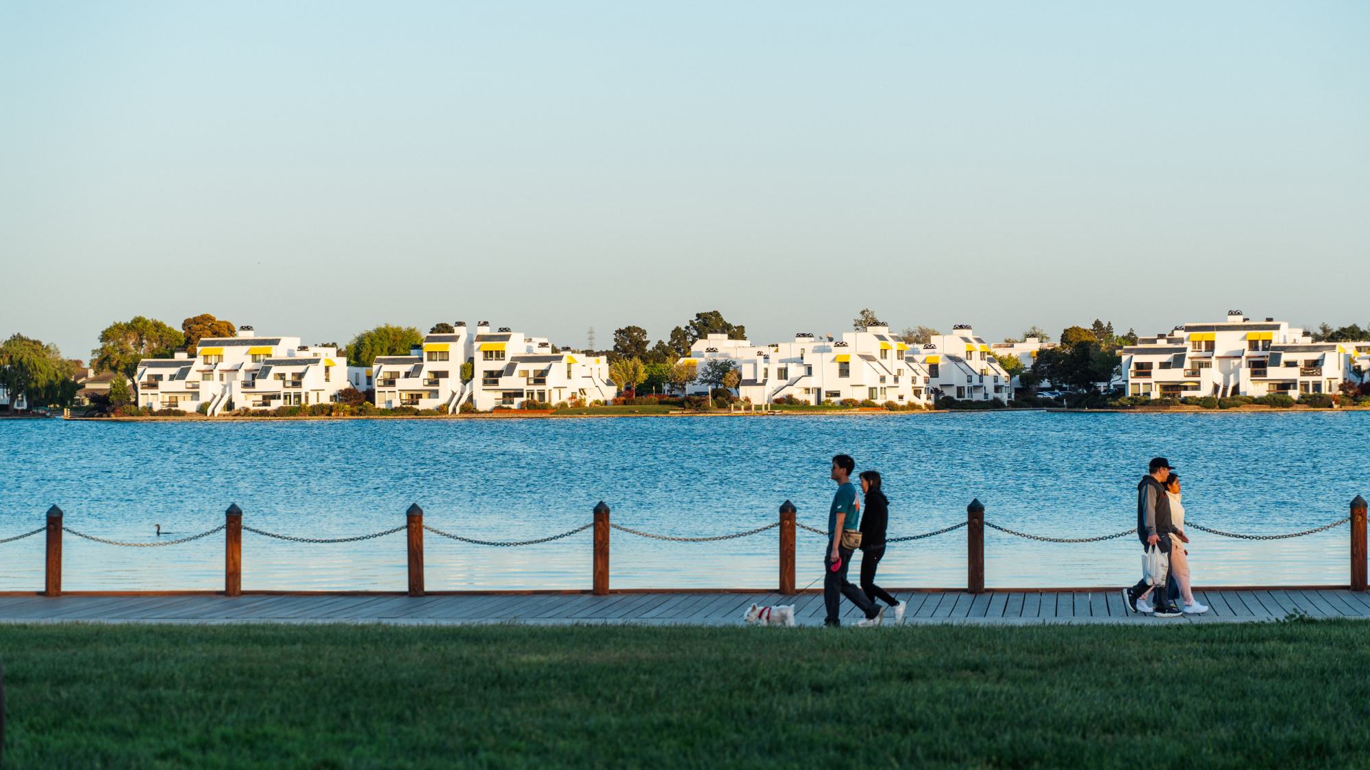 People walking along a waterfront boardwalk in Foster City with modern white homes across the lagoon in the background.