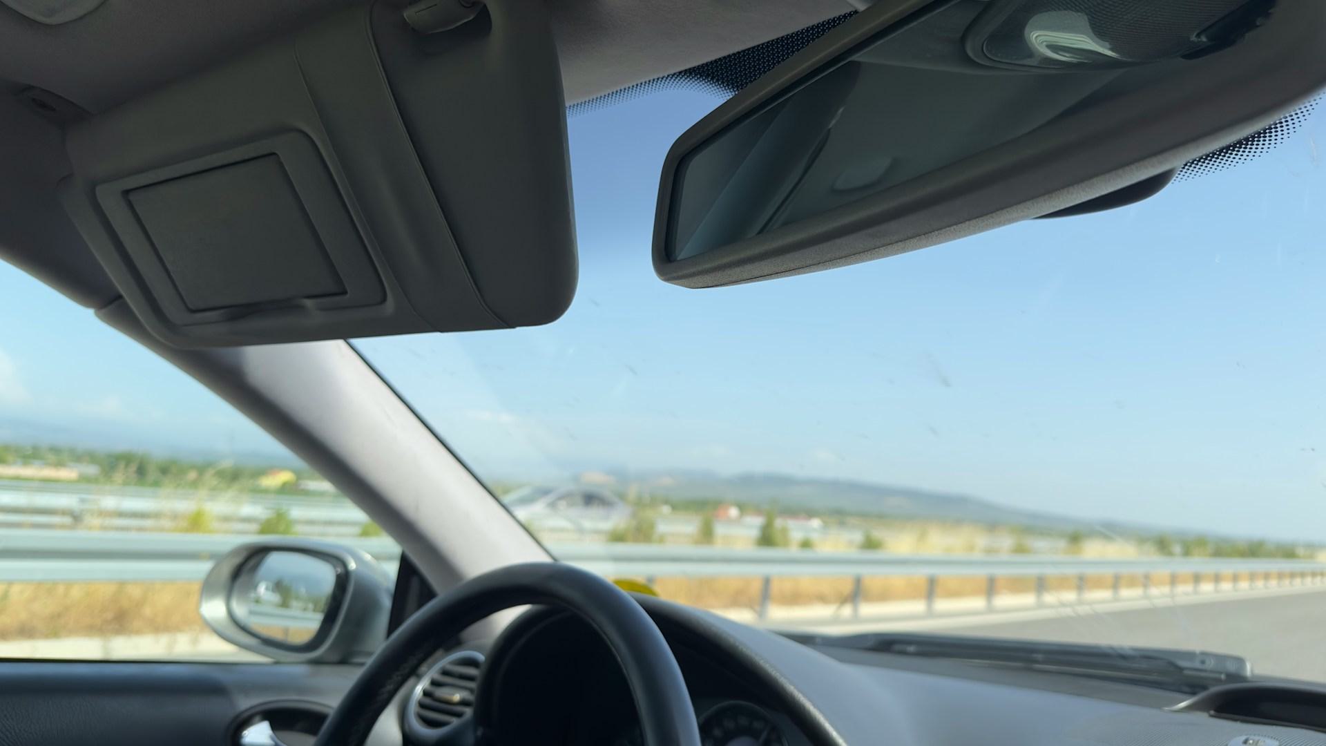 View from inside a car driving on a highway, showing the steering wheel, side mirror, and windshield with guardrails and distant hills visible under a clear sky.