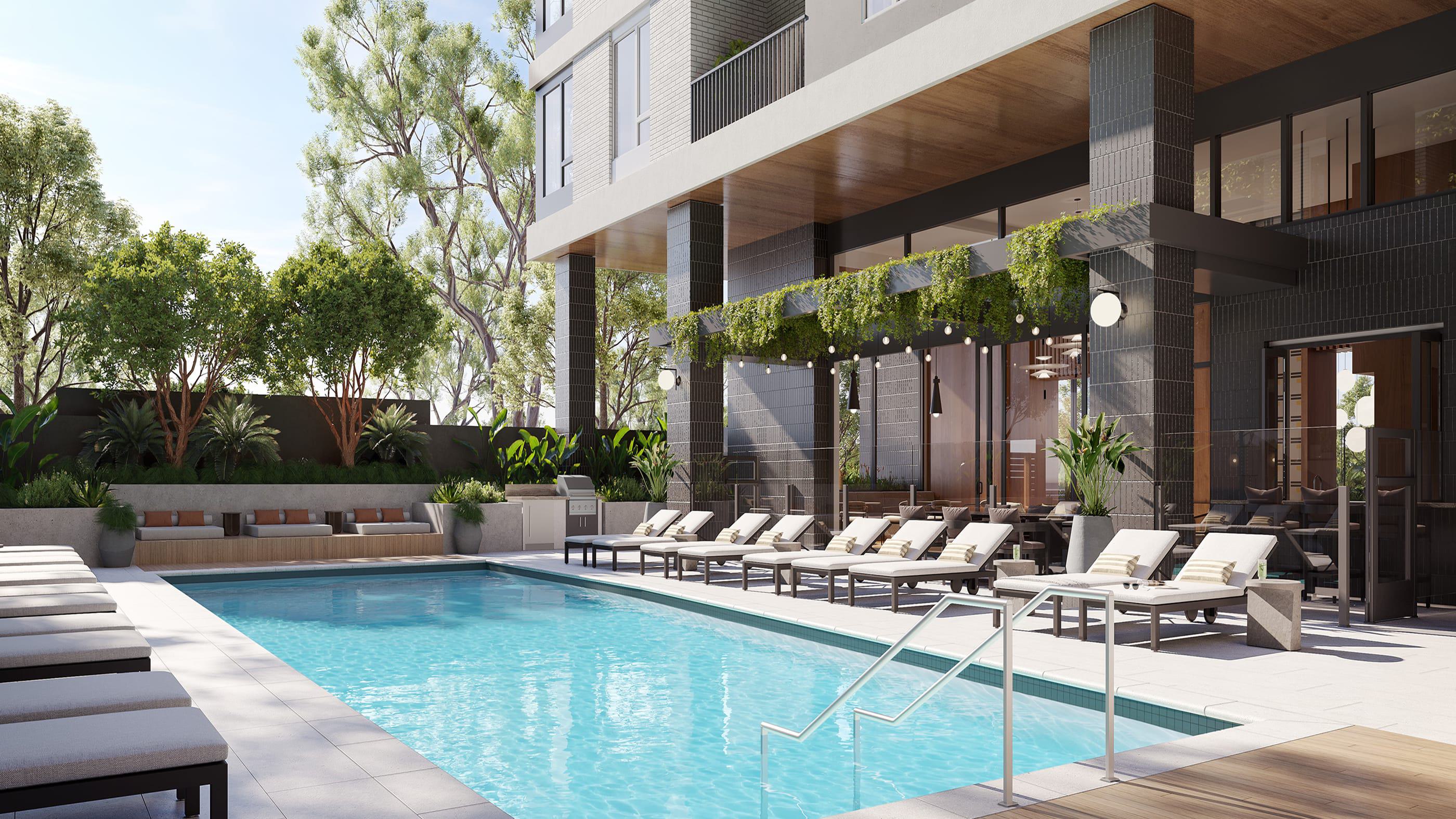 Outdoor pool deck at Revery with a rectangular swimming pool, rows of cushioned lounge chairs, built-in bench seating with pillows set against a landscaped planter, and a shaded area under the building overhang with tables, chairs, and hanging greenery.