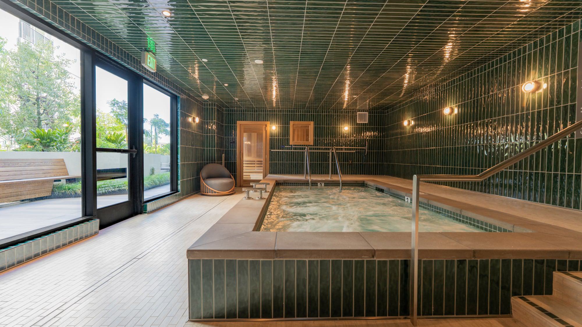 Indoor spa at Revery Apartments in Burlingame featuring a tiled soaking pool, warm wood accents, built-in bench seating, and soft lighting, with large windows opening to landscaped outdoor space