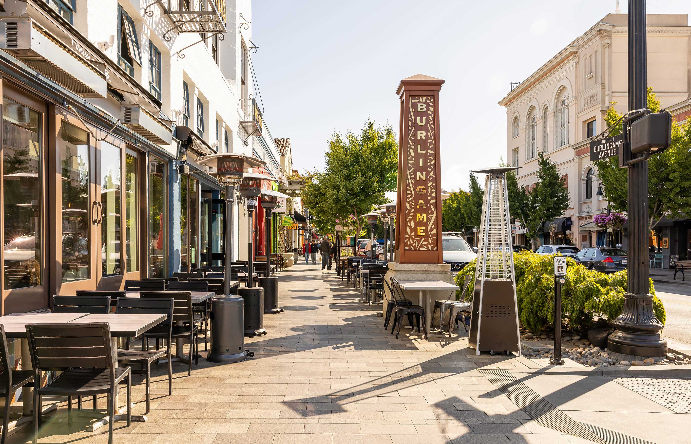 Street view of Burlingame Avenue with outdoor dining tables, shops, and a tall decorative sign reading Burlingame under tree-lined sidewalks.