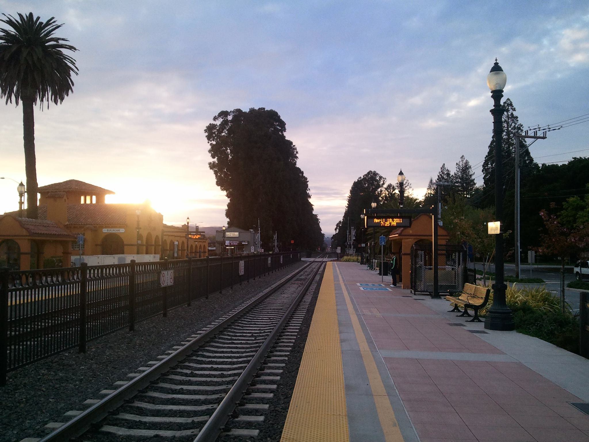 Burlingame Caltrain Station platform at sunrise with Spanish Revival–style station building, palm trees, benches, and digital departure sign