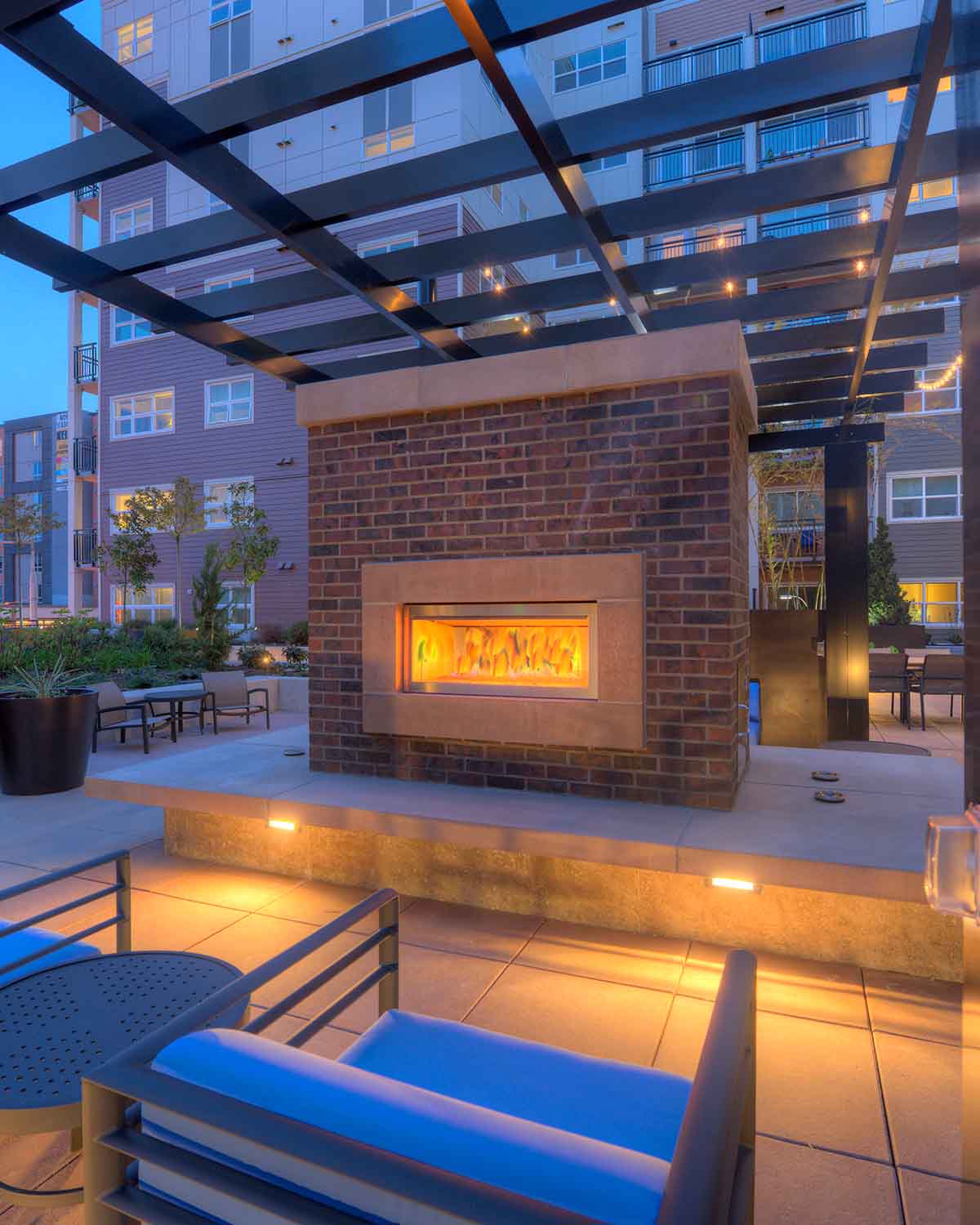 Outdoor courtyard lounge featuring a cozy brick fireplace, pergola overhead, and modern seating under warm evening lighting.