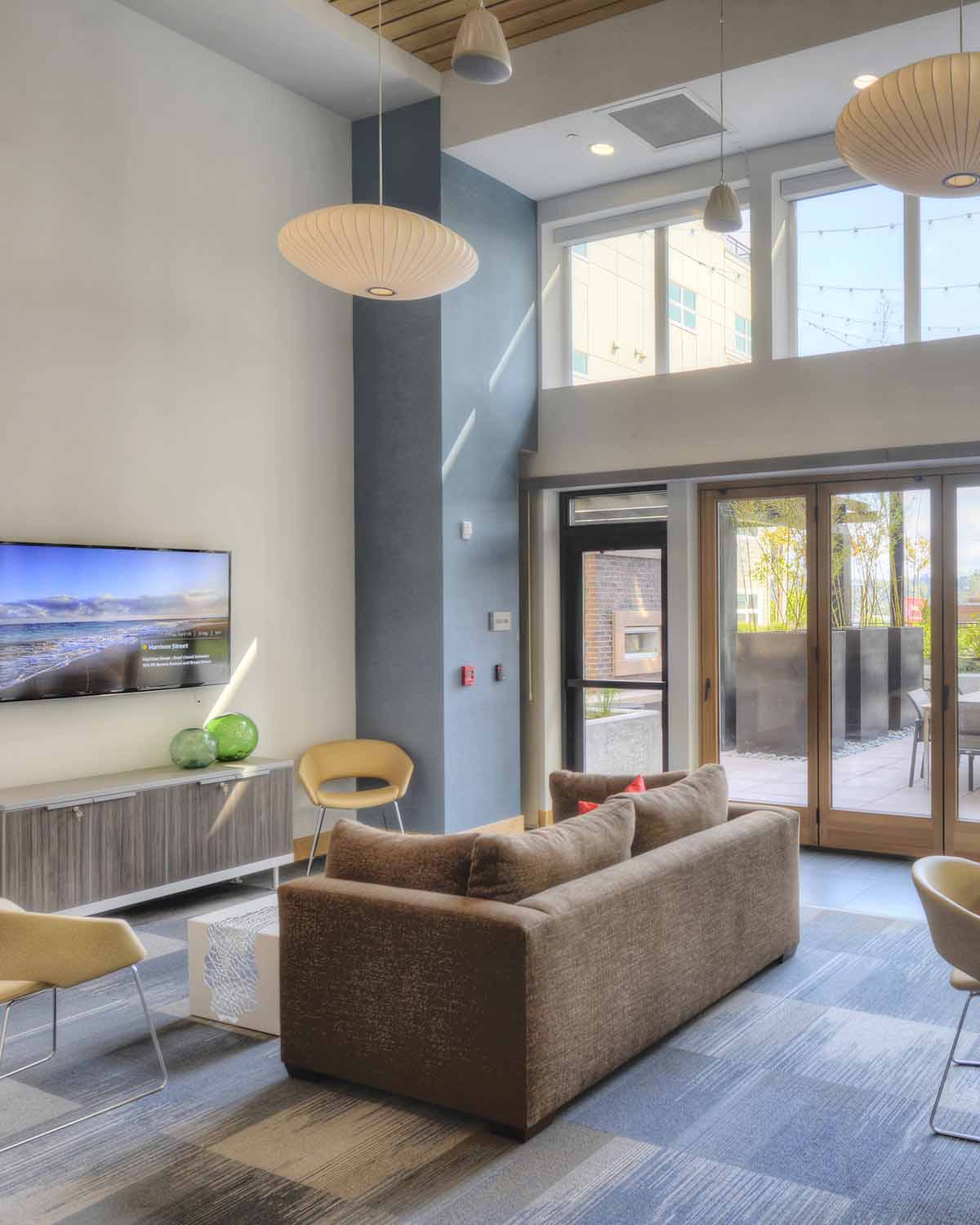 Modern resident lounge with a high ceiling, cozy seating, a wall-mounted TV, and access to an outdoor patio.