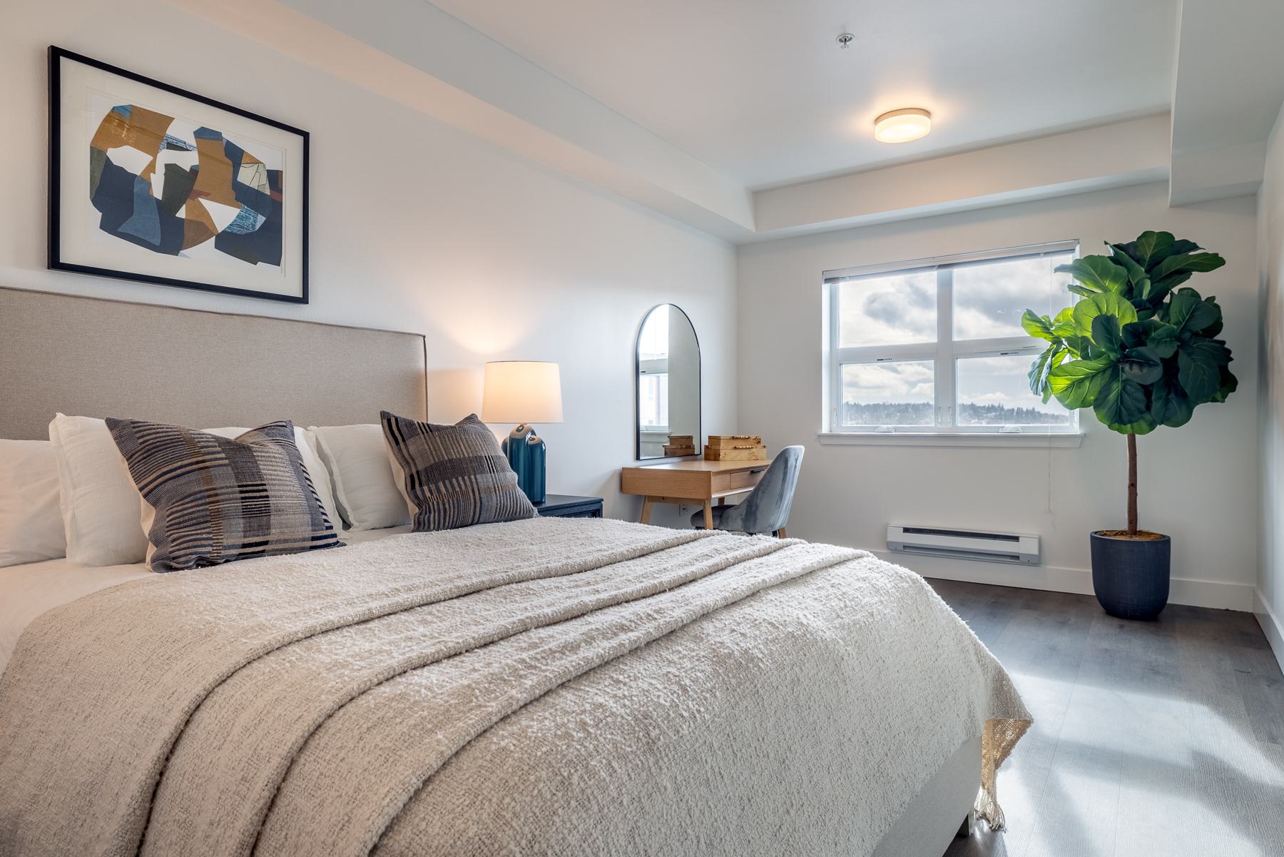 Cozy bedroom with neutral plaid bedding, a potted plant, and a large window offering natural light and views of a green hillside in Ballard in the distance.