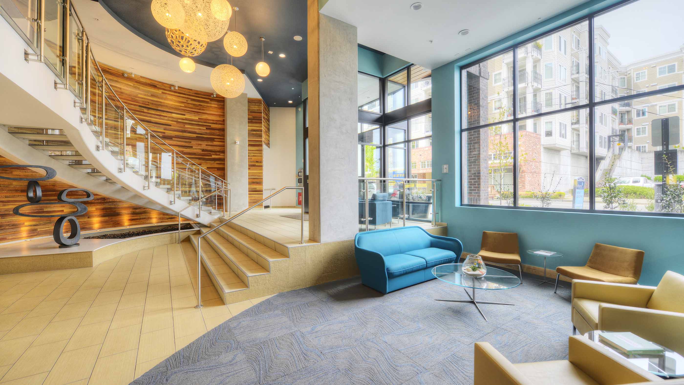 A modern apartment lobby in Ballard, WA, featuring a curved staircase, wood accent wall, pendant lights, and cozy seating by large street-facing windows